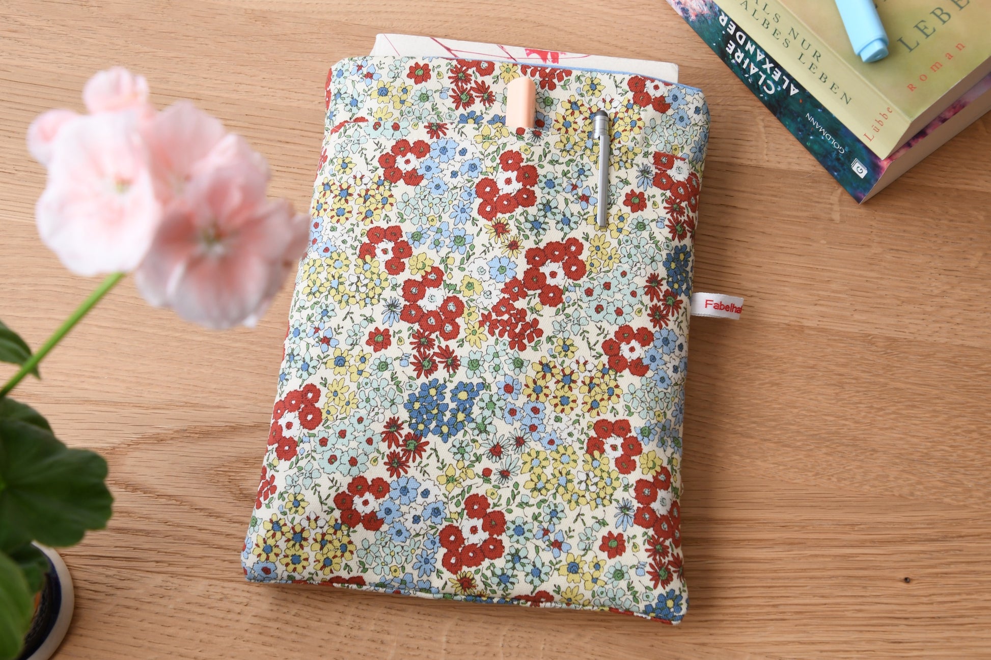 Handgefertigte, weich gepolsterte Buchtasche der Fabelhaften Werkstatt aus hellem Stoff mit einem zarten, detailreichen Streublumen-Muster in Pastelltönen. Gut sichtbar ist das aufgesetzte Vorderfach für Zubehör und die hochwertige Verarbeitung.