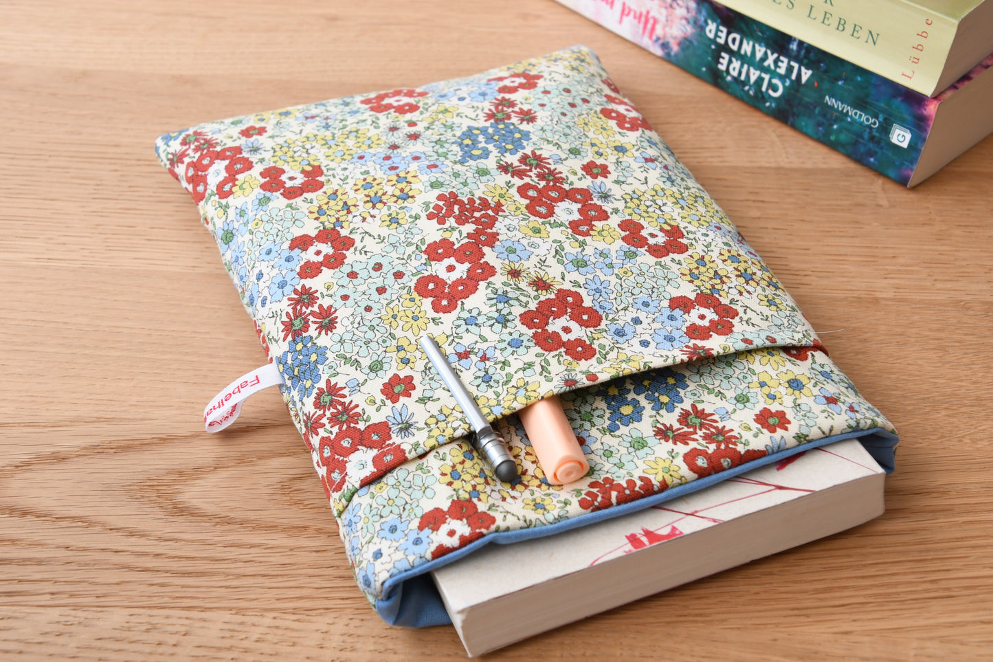  Buchtasche mit einem zarten, bunten Streublumen-Muster auf hellem Stoff. Ein Taschenbuch schaut oben ein Stück aus der weich gepolsterten Hülle heraus. Im vorderen Fach stecken ordentlich zwei Stifte für die nächste Lesepause.