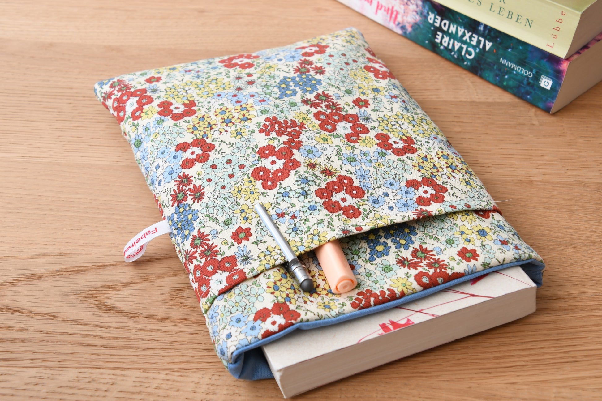  Buchtasche mit einem zarten, bunten Streublumen-Muster auf hellem Stoff. Ein Taschenbuch schaut oben ein Stück aus der weich gepolsterten Hülle heraus. Im vorderen Fach stecken ordentlich zwei Stifte für die nächste Lesepause.