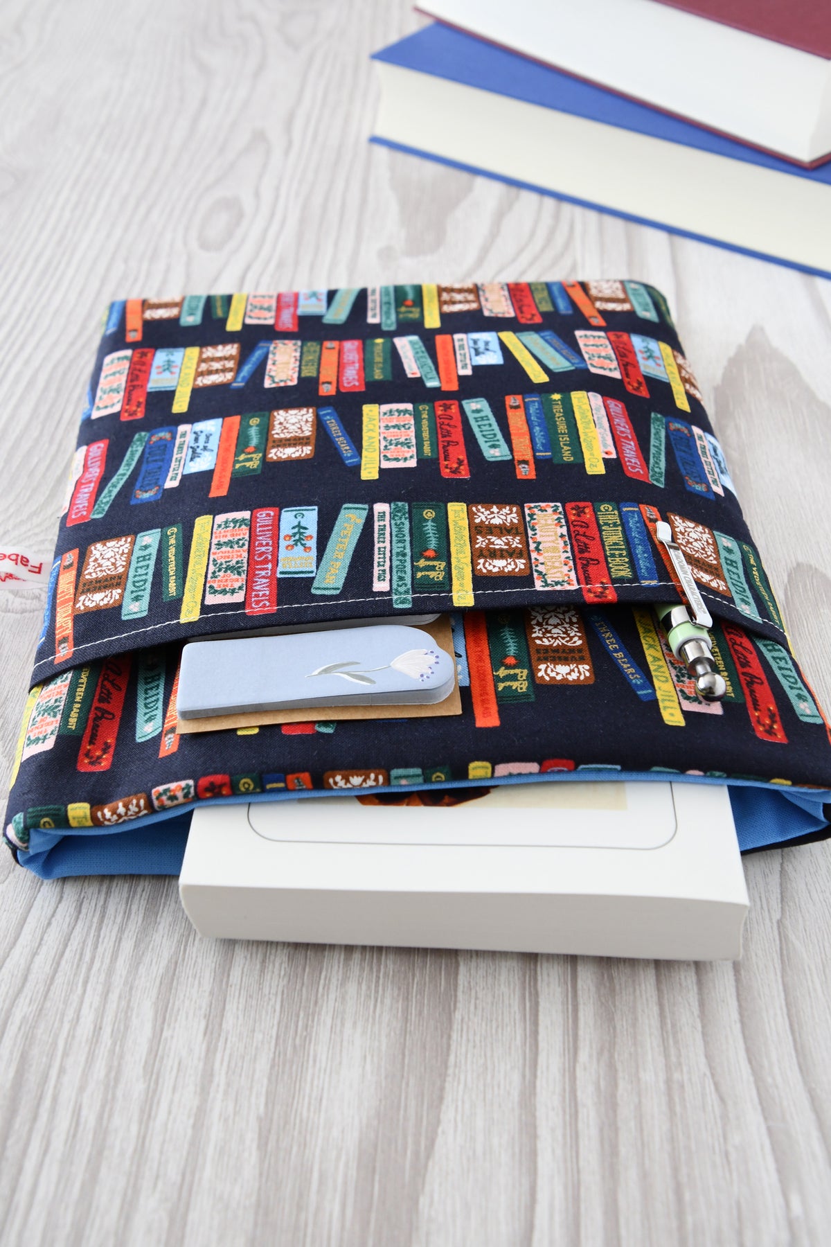 Detailreiches Foto einer Buchtasche mit Bücherrücken-Print. Aus der weichen Hülle schaut ein echtes Buch heraus. Das clevere Zubehörfach auf der Vorderseite ist mit einem Kugelschreiber bestückt, um den perfekten Schutz für unterwegs zu demonstrieren.