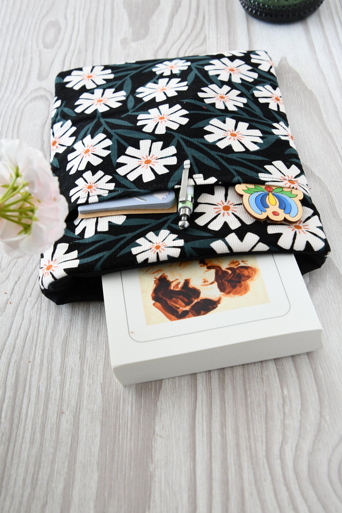 Fröhliche, weich gepolsterte Buchtasche mit einem sommerlichen Margeriten-Muster (weiße Blüten mit orangenfarbender Mitte). Aus der oberen Öffnung ragt ein Roman heraus, und im Vorderfach sind Stifte und kleine Haftnotizen griffbereit platziert.