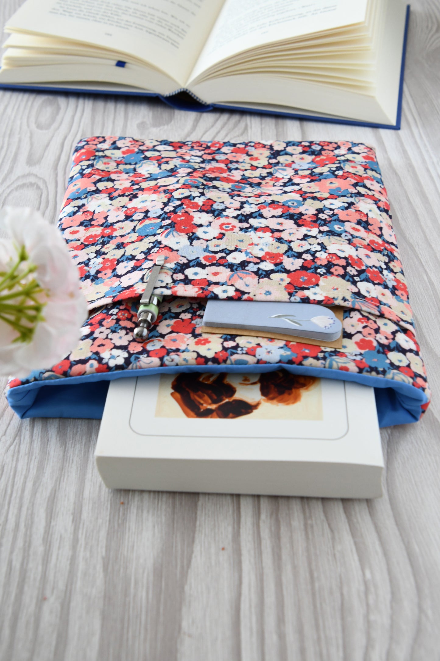 Draufsicht auf die geöffnete Buchtasche Jenifer im Blumenkonfetti-Design mit eingestecktem Buch sowie Stift und Lesezeichen in der Vordertasche.
