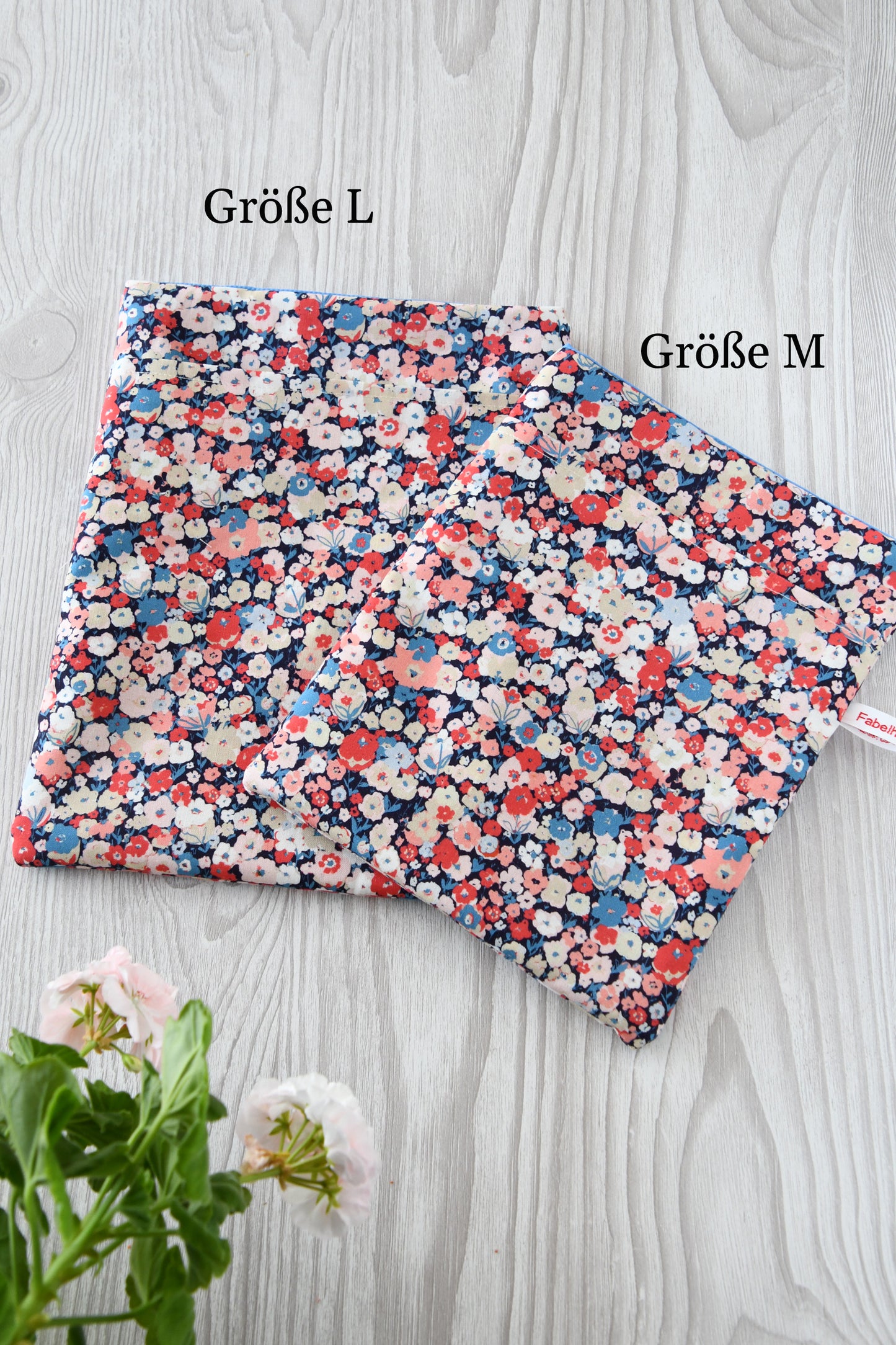 Handgemachte Buchtaschen aus Stoff im Größenvergleich nebeneinander: Die kleine Buchhülle für Taschenbücher und eReader liegt links neben der großen Schutzhülle für gebundene Bücher und 10-Zoll-Tablets. Beide Taschen der Fabelhaften Werkstatt Bremen zeigen das bunte Blumenkonfetti-Muster "Jenifer" und sind weich gepolstert.