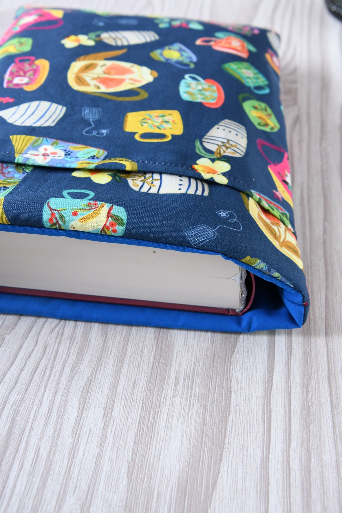 Große Buchtasche „Teatime“ in Größe L mit Teetassen-Muster auf blauem Grund. Ein dicker Hardcover-Wälzer (ca. 500 Seiten) ist passgenau in die gepolsterte Buchhülle eingesteckt.