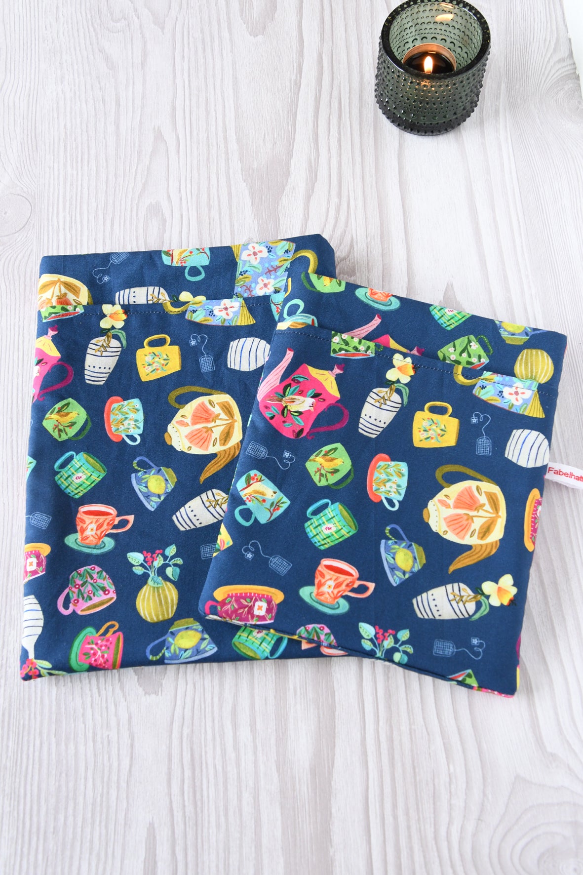 Größenvergleich der Buchtasche „Teatime“ im bunten Teetassen-Design: Links Größe L für dicke Hardcover-Bücher und Tablets, rechts Standardgröße M für Taschenbücher, nebeneinander arrangiert.