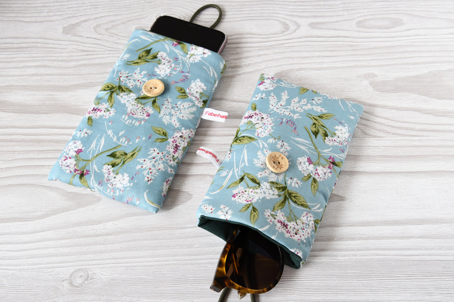 Zwei handgenähte Stofftaschen von Fabelhafte Werkstatt nebeneinander: Eine genutzt als schützende Handyhülle mit Smartphone, die andere als leichtes Brillenetui mit einer Sonnenbrille.