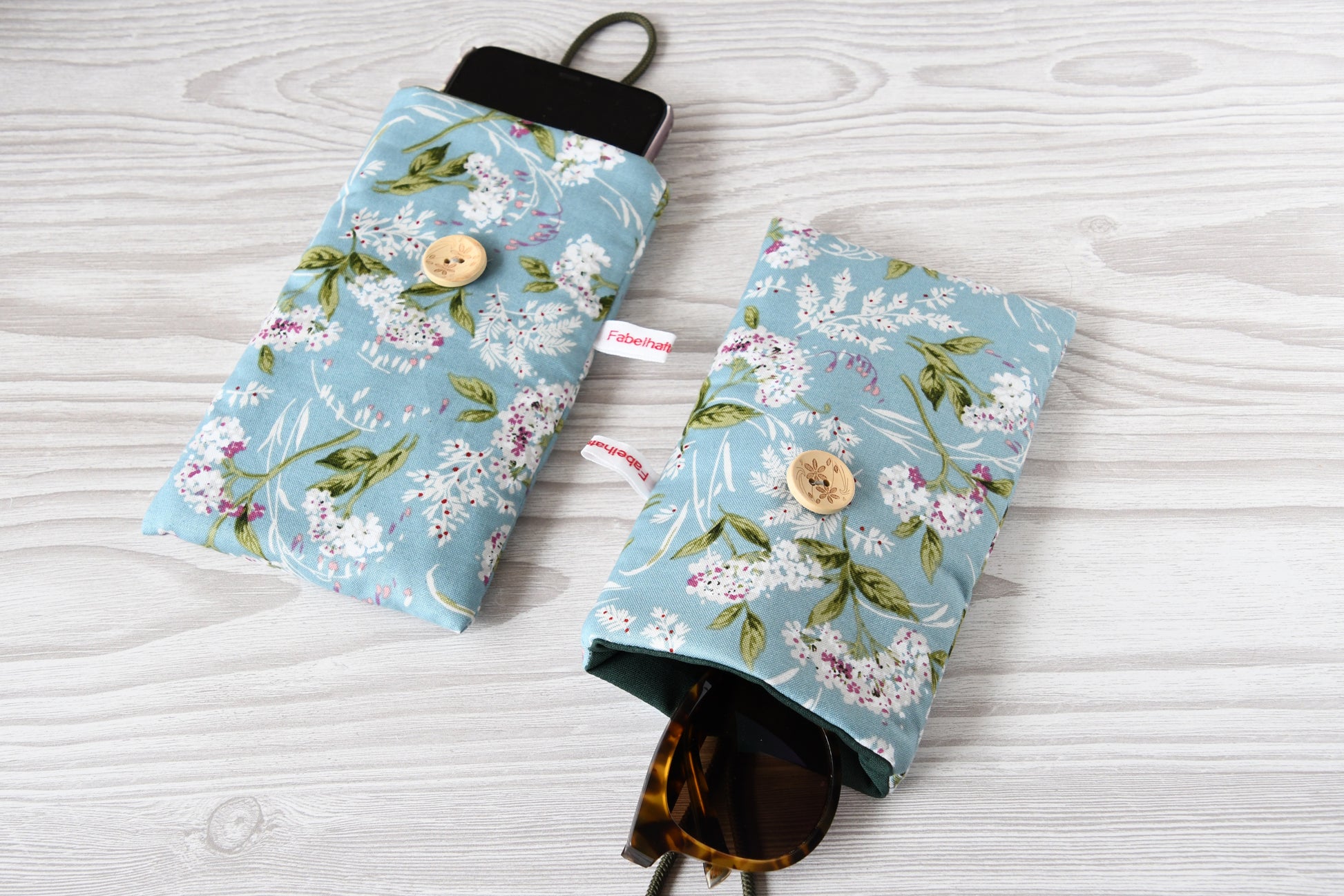 Zwei handgenähte Stofftaschen von Fabelhafte Werkstatt nebeneinander: Eine genutzt als schützende Handyhülle mit Smartphone, die andere als leichtes Brillenetui mit einer Sonnenbrille.