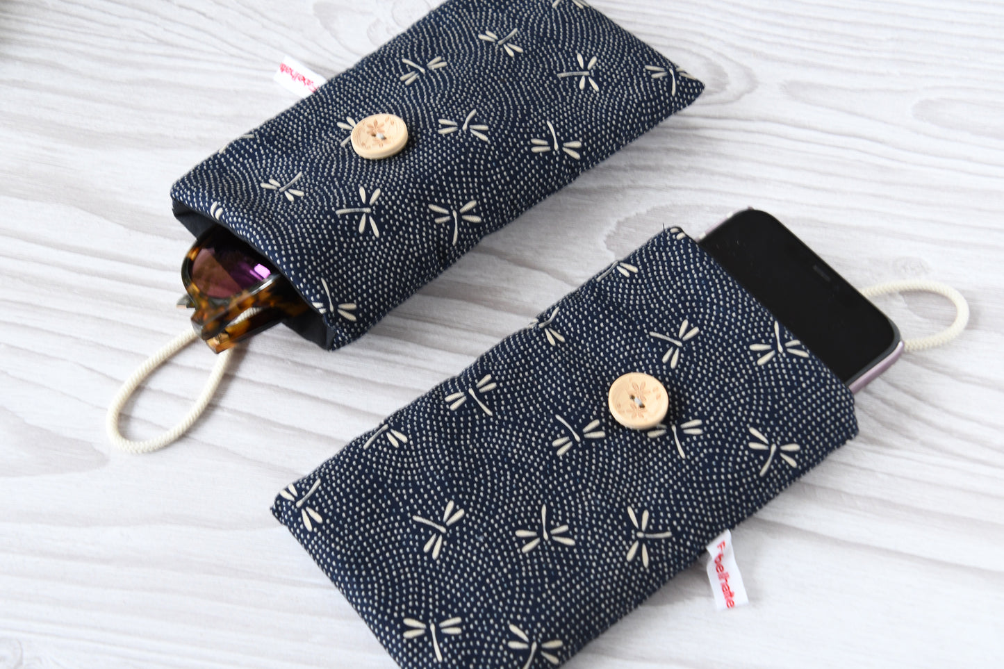 Zwei handgenähte Stofftaschen von Fabelhafte Werkstatt nebeneinander: Eine genutzt als schützende Handyhülle mit Smartphone, die andere als leichtes Brillenetui mit einer Sonnenbrille.