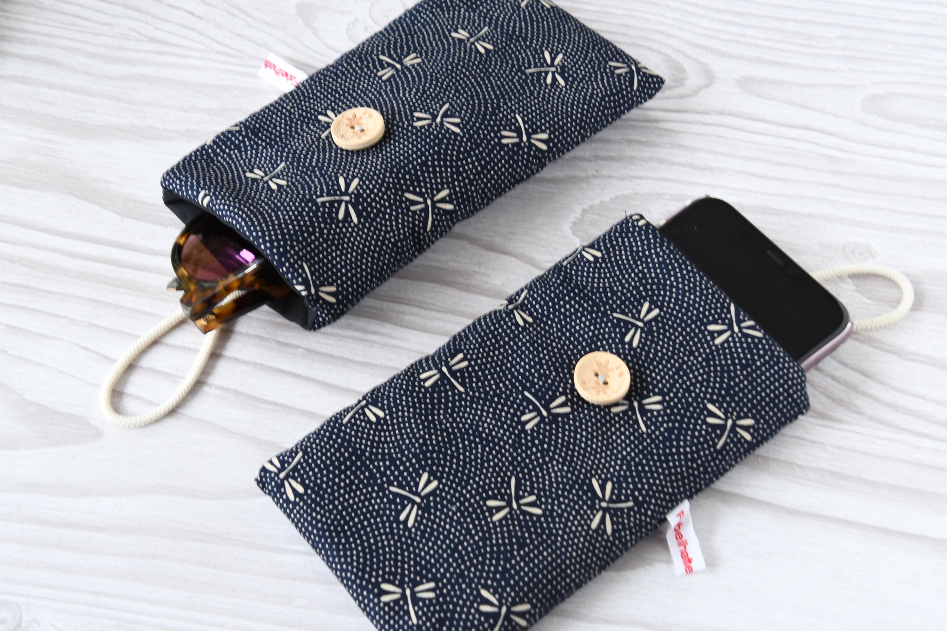 Zwei handgenähte Stofftaschen von Fabelhafte Werkstatt nebeneinander: Eine genutzt als schützende Handyhülle mit Smartphone, die andere als leichtes Brillenetui mit einer Sonnenbrille.