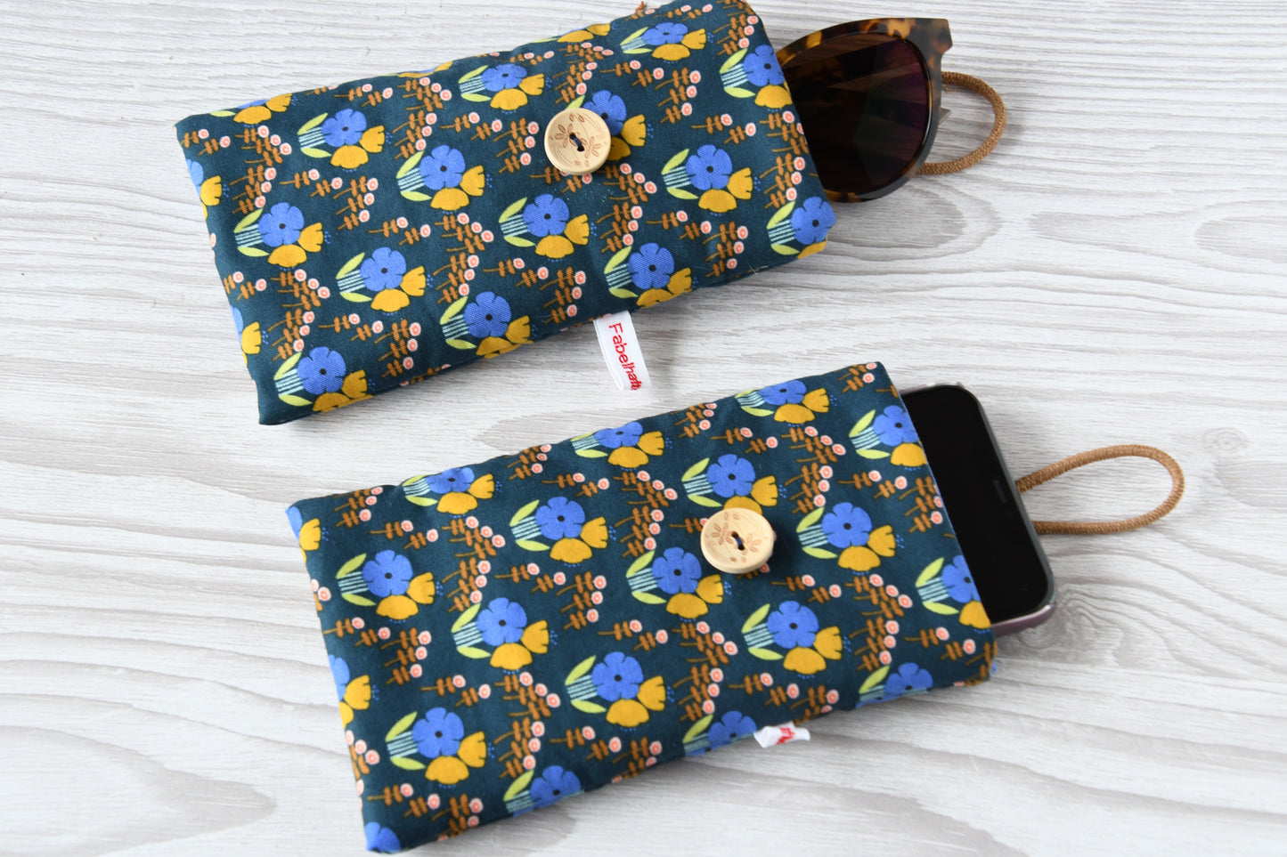 Zwei handgenähte Stofftaschen von Fabelhafte Werkstatt nebeneinander: Eine genutzt als schützende Handyhülle mit Smartphone, die andere als leichtes Brillenetui mit einer Sonnenbrille.