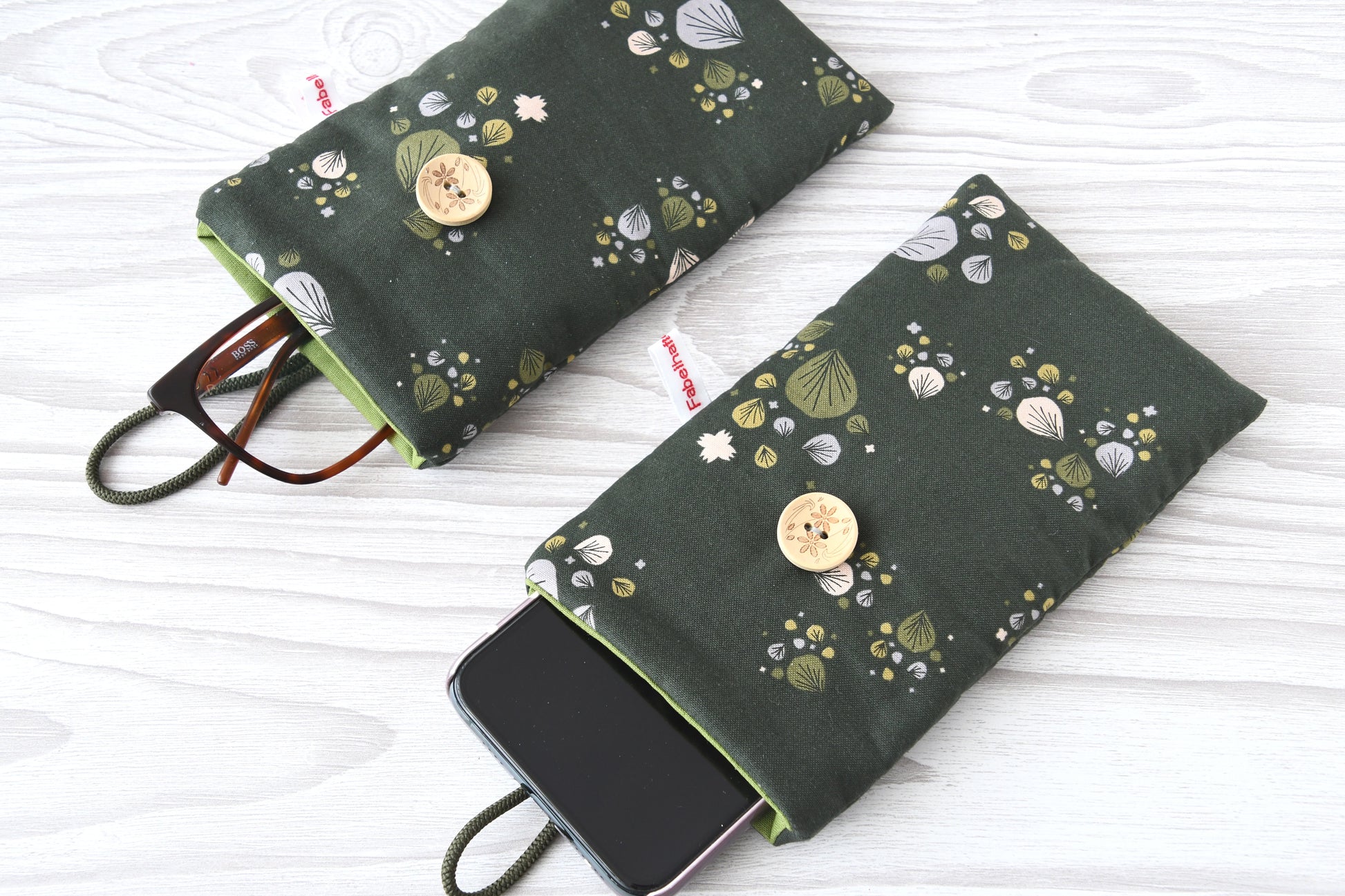 Zwei handgenähte Stofftaschen von Fabelhafte Werkstatt nebeneinander: Eine genutzt als schützende Handyhülle mit Smartphone, die andere als leichtes Brillenetui mit einer Sonnenbrille.