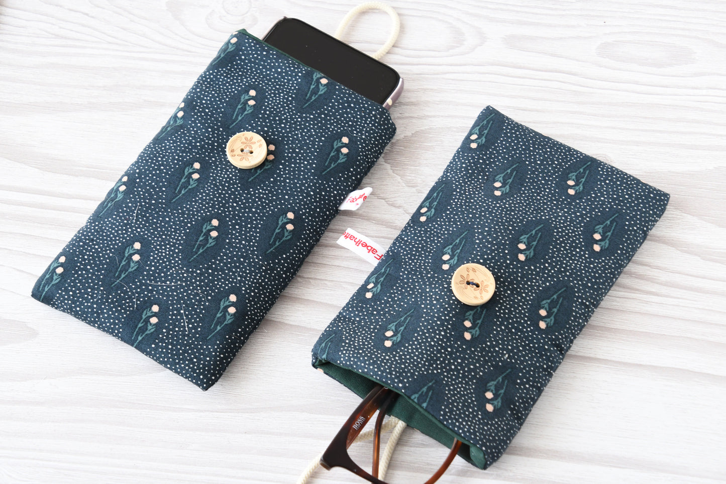 Zwei handgenähte Stofftaschen von Fabelhafte Werkstatt nebeneinander: Eine genutzt als schützende Handyhülle mit Smartphone, die andere als leichtes Brillenetui mit einer Sonnenbrille.