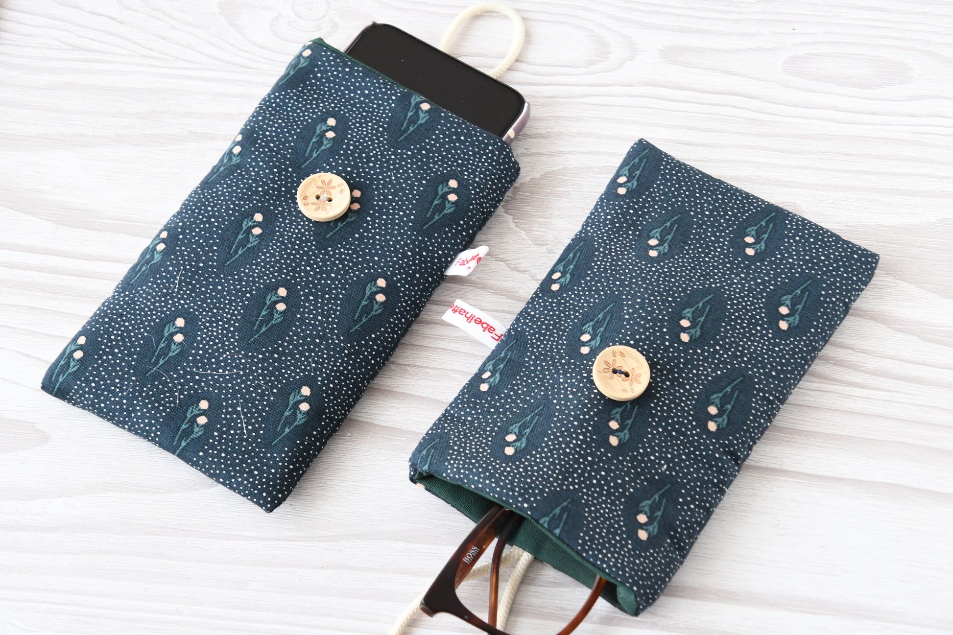 Zwei handgenähte Stofftaschen von Fabelhafte Werkstatt nebeneinander: Eine genutzt als schützende Handyhülle mit Smartphone, die andere als leichtes Brillenetui mit einer Sonnenbrille.