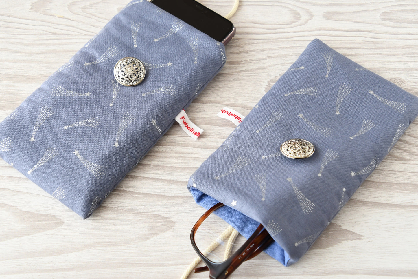 Zwei handgenähte Stofftaschen mit silbernen Sternschnuppen von Fabelhafte Werkstatt nebeneinander: Eine genutzt als Handyhülle mit Smartphone, die andere als Brillenetui mit Sonnenbrille.
