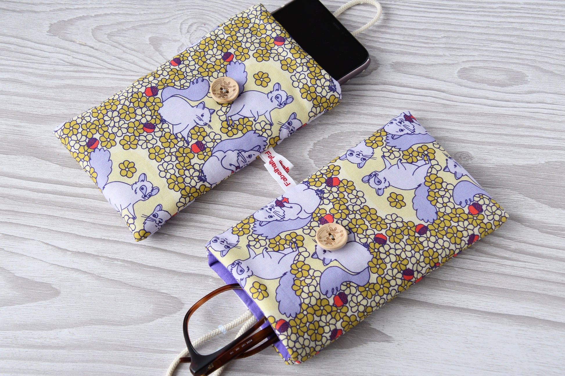 Zwei handgenähte Stofftaschen mit süßem Eichhörnchen-Muster von Fabelhafte Werkstatt nebeneinander: Eine genutzt als Handyhülle mit Smartphone, die andere als Brillenetui mit Sonnenbrille.