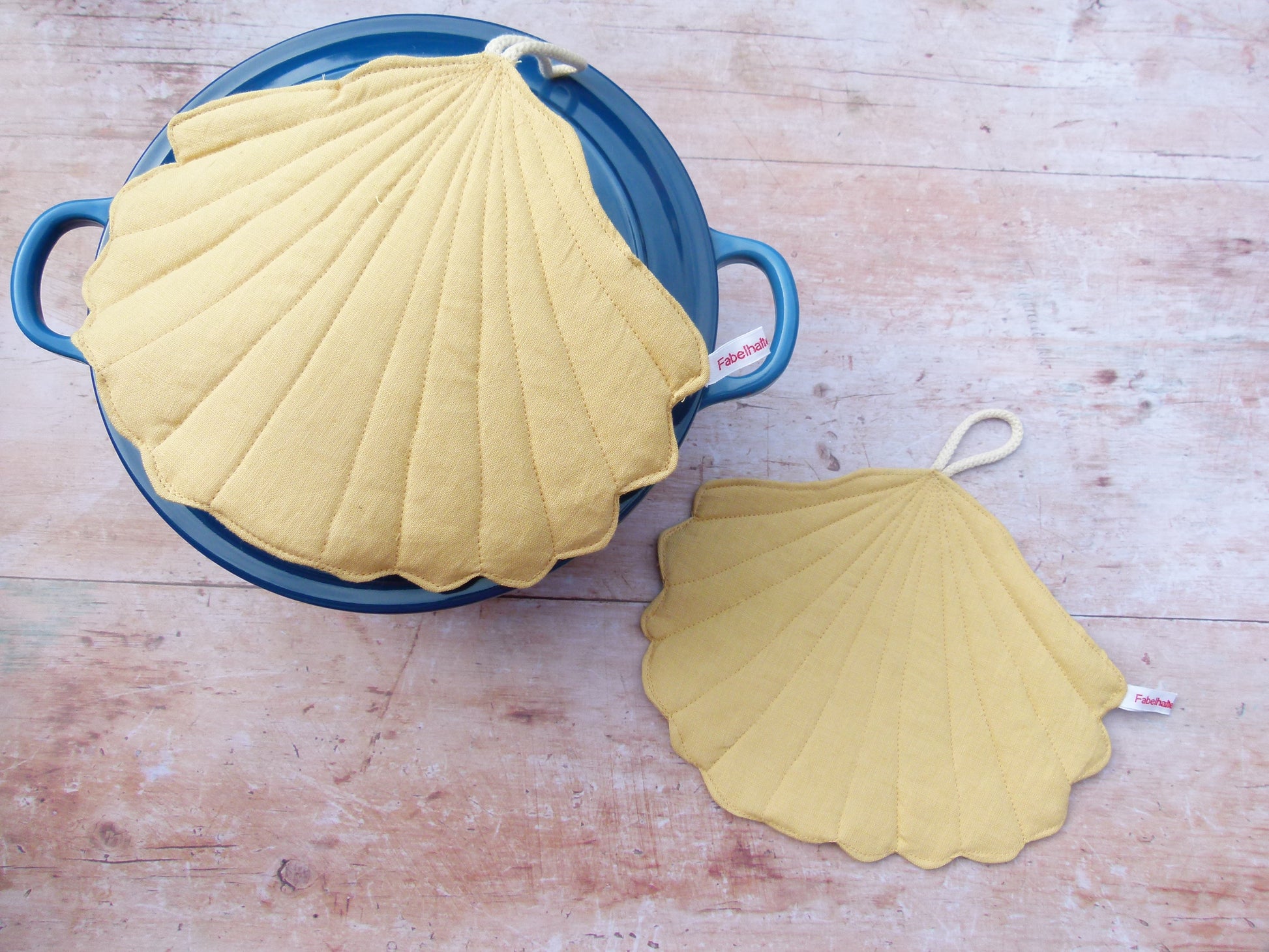 Präsentation des maritimen Topflappen-Sets an einem Kochtopf. Ein muschelförmiger Topflappen aus gelbem Leinen liegt flach auf dem Topfdeckel, während der zweite Topflappen dekorativ daneben auf der Küchenzeile platziert ist.