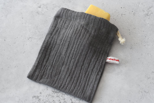 Handgenähtes Seifensäckchen von Fabelhaftewerkstatt aus grauem Baumwoll-Musselin. Ein Stück helle Naturseife schaut ein Stück weit aus der geöffneten Öffnung des weichen Stoffbeutels heraus.