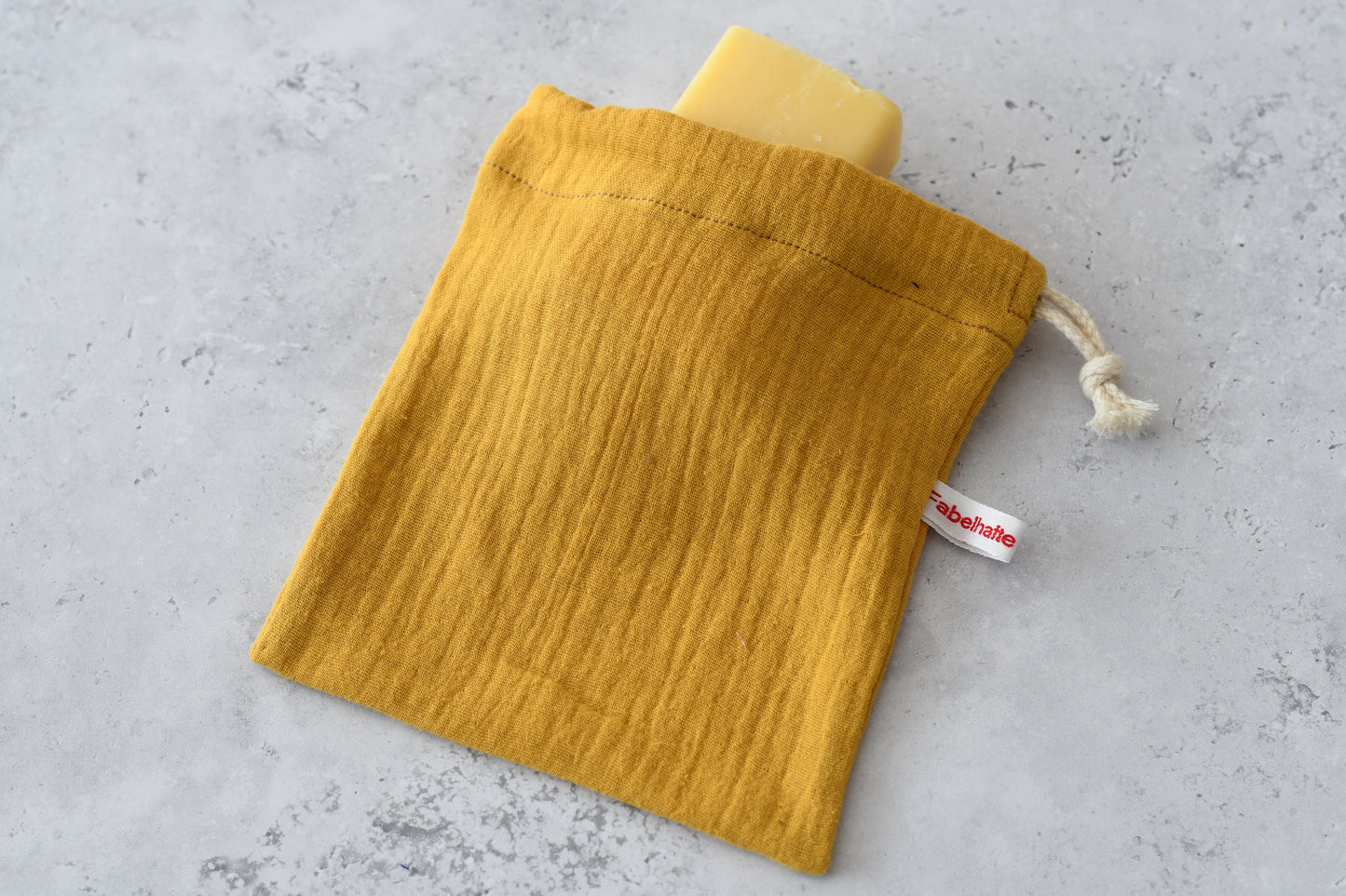 Zero Waste im Badezimmer: Handgemachter Seifenbeutel aus senfgelben Musselin mit Naturseife auf einer hellen Unterlage für die plastikfreie Körperpflege.
