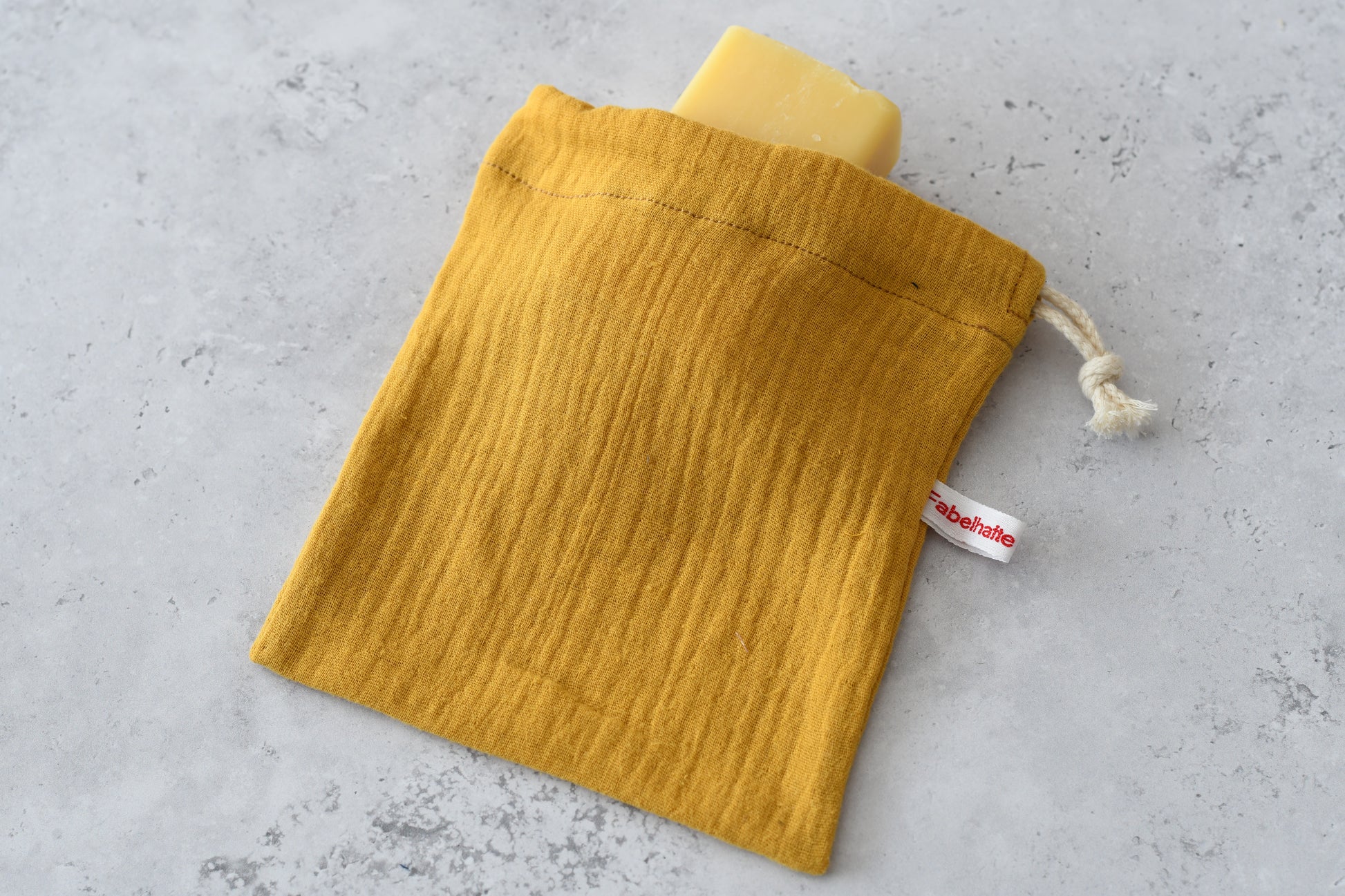 Zero Waste im Badezimmer: Handgemachter Seifenbeutel aus senfgelben Musselin mit Naturseife auf einer hellen Unterlage für die plastikfreie Körperpflege.