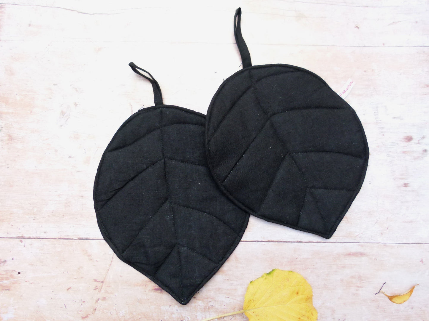 Zwei blattförmige Topflappen aus schwarzen Leinen im Set () liegen flach nebeneinander auf einer hellen Küchenarbeitsplatte.