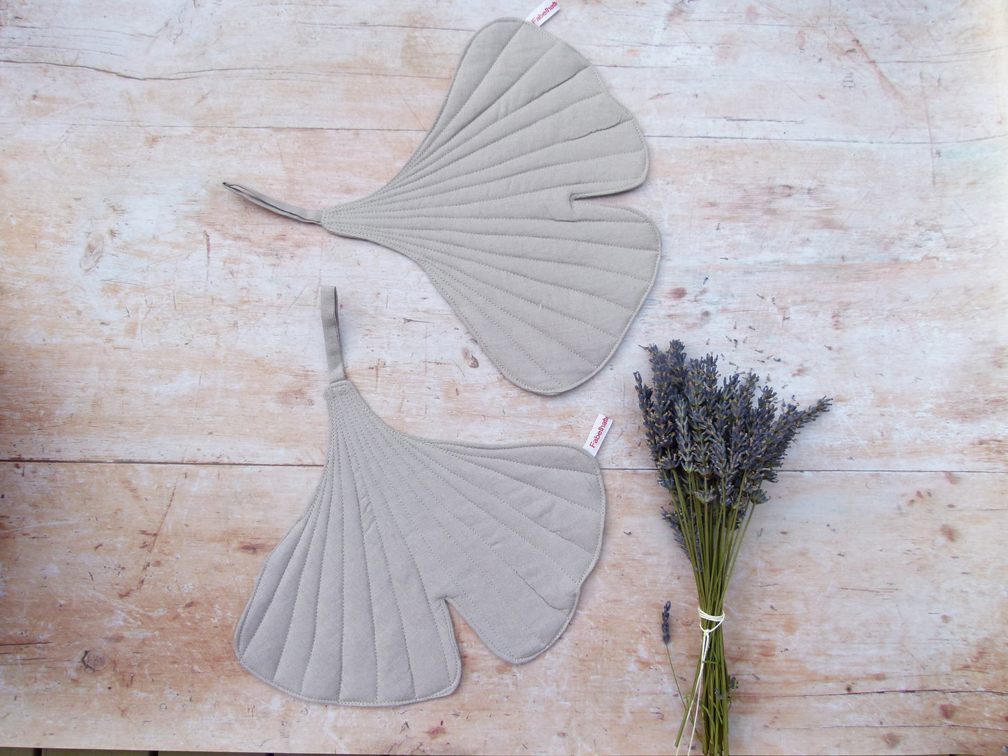 Ein Set aus zwei handgenähten Topflappen in der markanten Form eines Ginkgoblattes aus reinem, hellgrauen Leinen, flach liegend auf einem Holztisch.