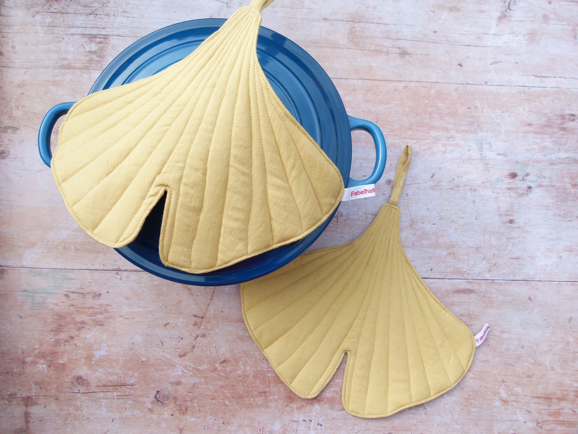 Ein Set aus zwei handgenähten Topflappen in der Form eines Ginkgoblattes, gefertigt aus gelben Leinen.  Topfpappen liegen auf dem Deckel eines dunklen Gusseisentopfes. Eine Aufhängeschlaufe und ein kleines Etikett sind sichtbar.
