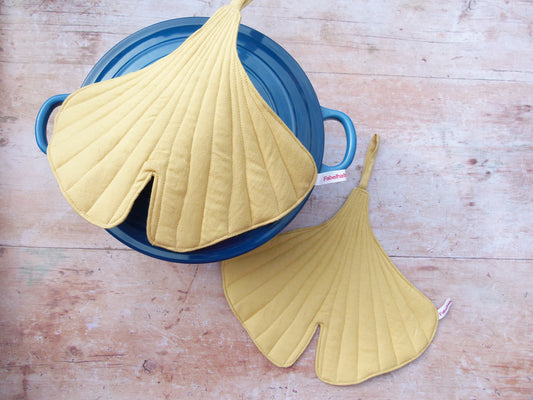 Ein Set aus zwei handgenähten Topflappen in der Form eines Ginkgoblattes, gefertigt aus gelben Leinen.  Topfpappen liegen auf dem Deckel eines dunklen Gusseisentopfes. Eine Aufhängeschlaufe und ein kleines Etikett sind sichtbar.