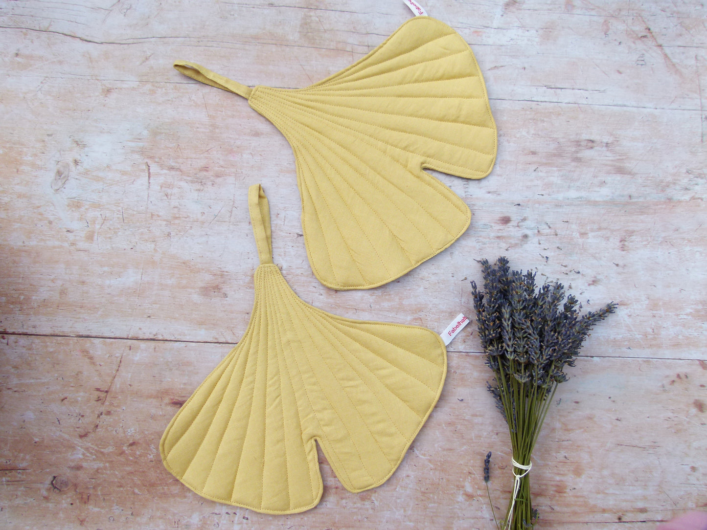 Ein Set aus zwei handgenähten Topflappen in der markanten Form eines Ginkgoblattes aus reinem, gelben Leinen, flach liegend auf einem Holztisch