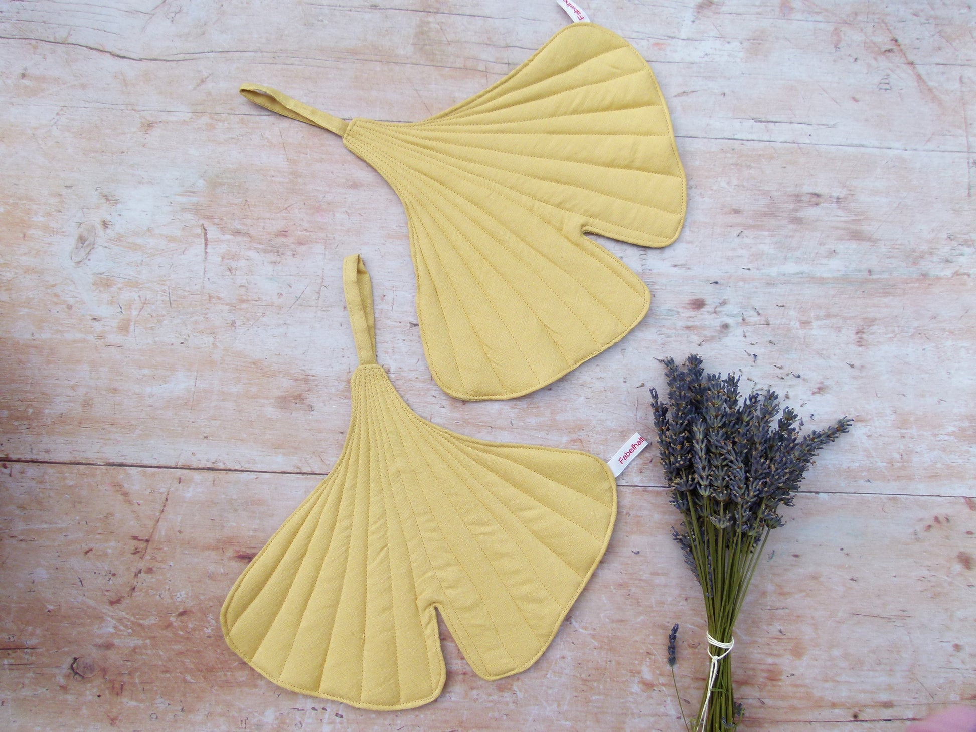Ein Set aus zwei handgenähten Topflappen in der markanten Form eines Ginkgoblattes aus reinem, gelben Leinen, flach liegend auf einem Holztisch