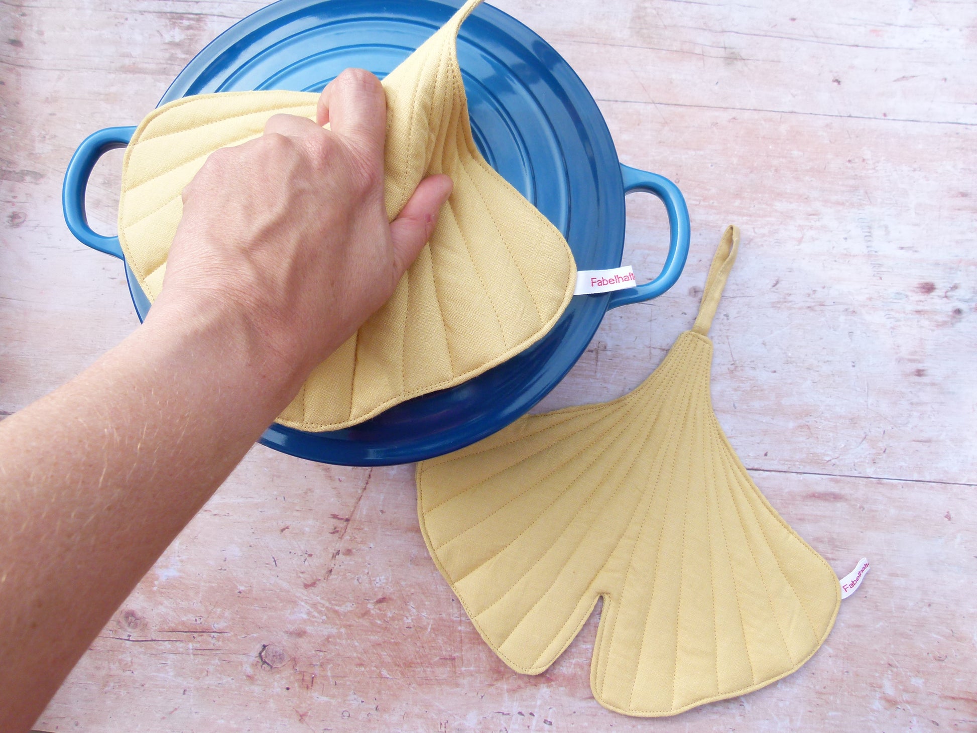 Ein Set aus zwei handgenähten Topflappen in der Form eines Ginkgoblattes, gefertigt aus gelben Leinen, auf einem  dunklen Gusseisentopf. Eine Aufhängeschlaufe und ein kleines Etikett sind sichtbar. 