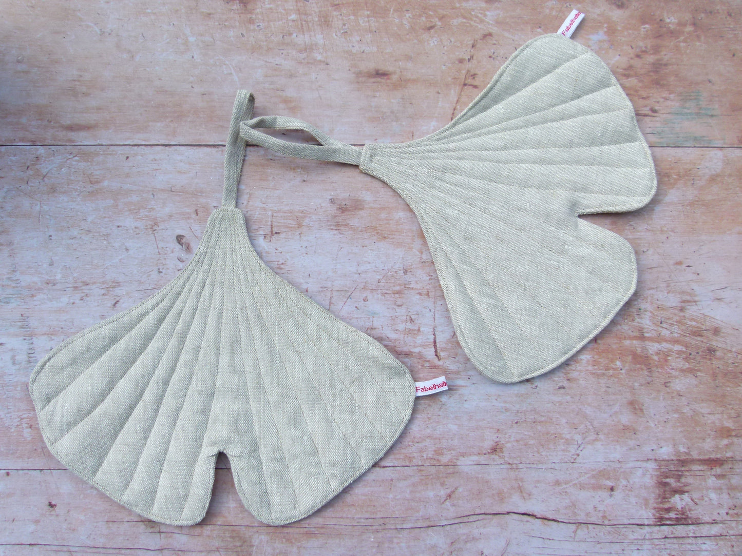 in Set aus zwei handgenähten Topflappen in der markanten Form eines Ginkgoblattes aus reinem, naturfarbenem Leinen, flach liegend auf einem Holztisch.