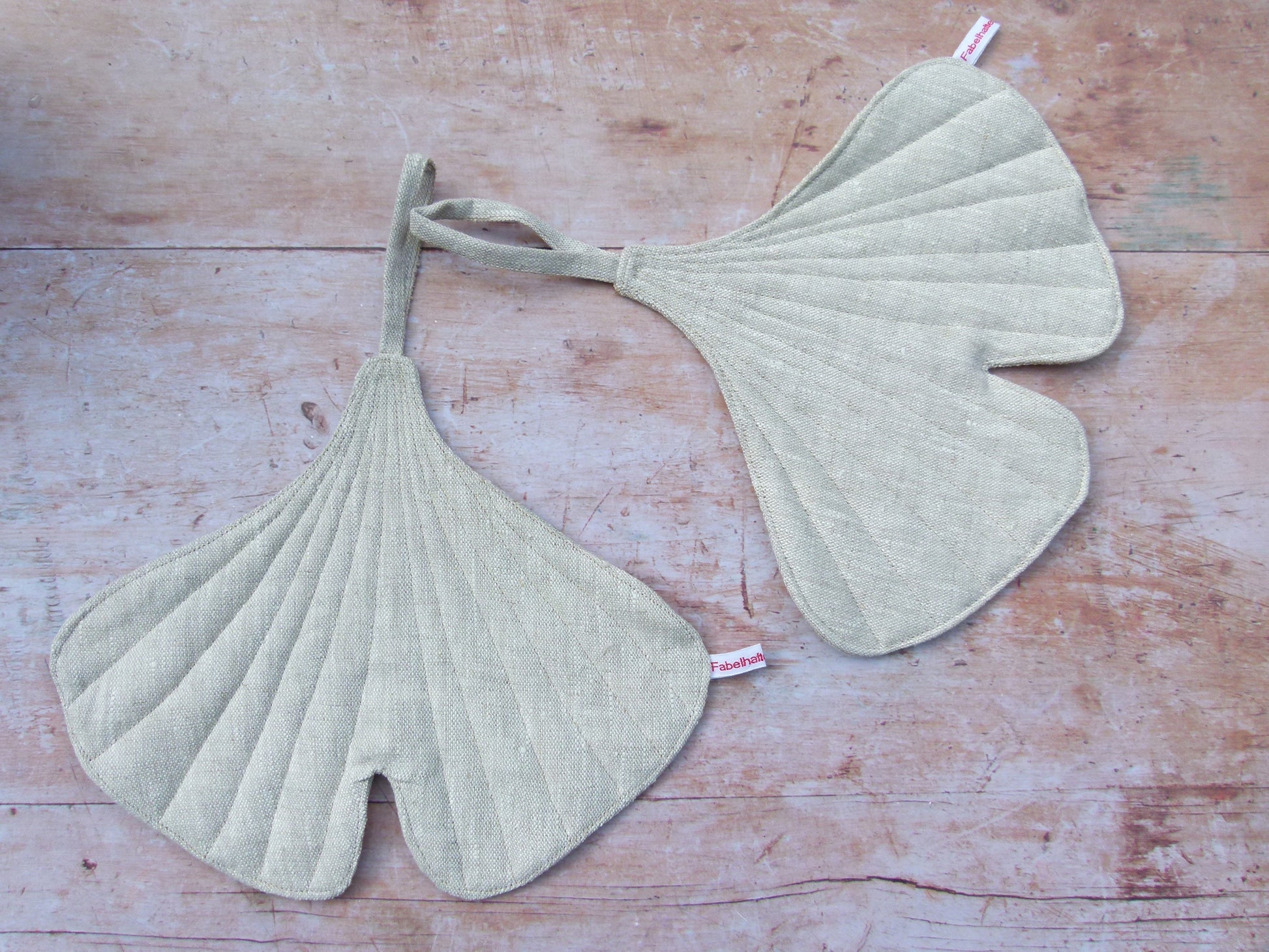 in Set aus zwei handgenähten Topflappen in der markanten Form eines Ginkgoblattes aus reinem, naturfarbenem Leinen, flach liegend auf einem Holztisch.