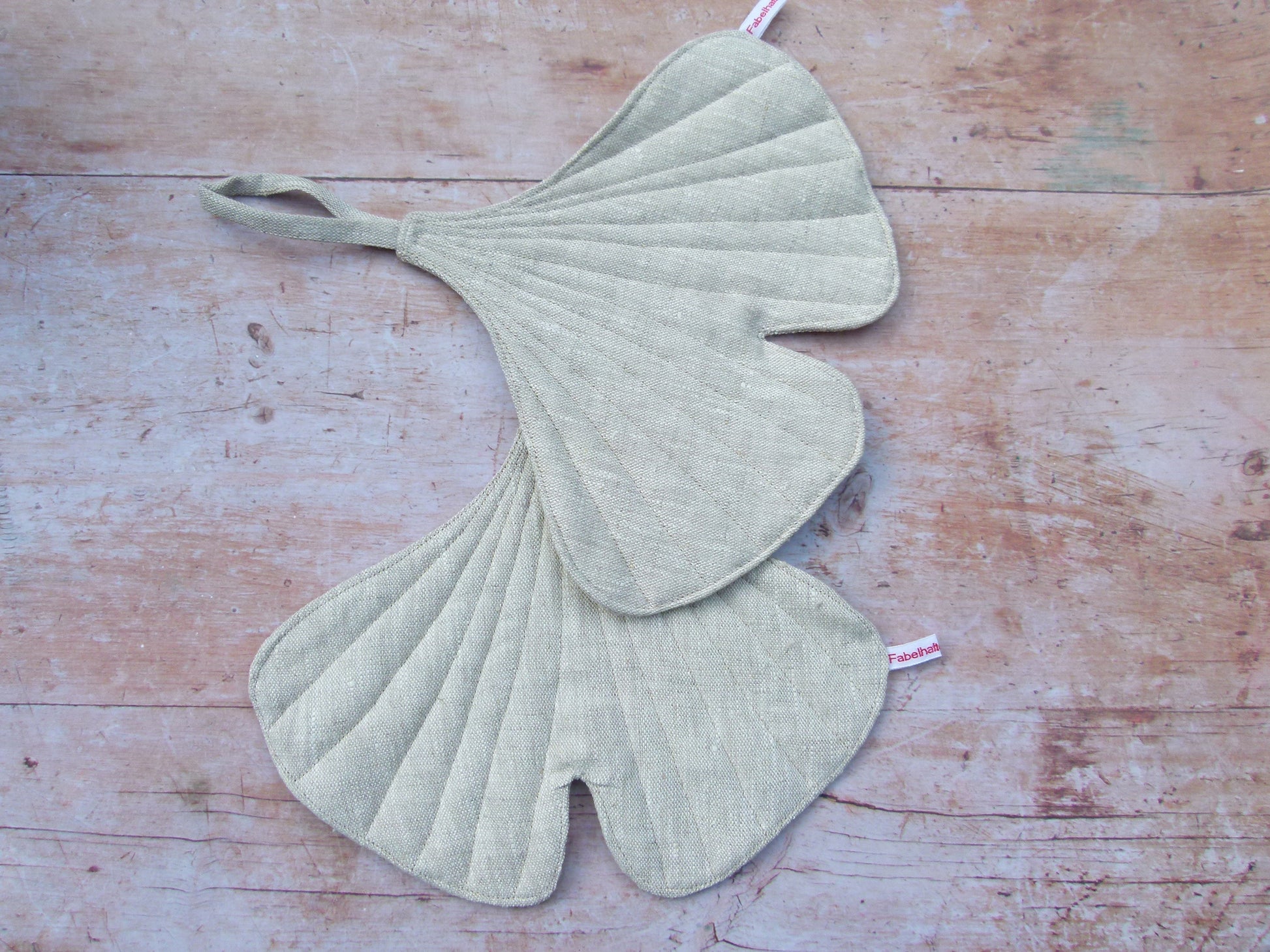 in Set aus zwei handgenähten Topflappen in der markanten Form eines Ginkgoblattes aus reinem, naturfarbenem Leinen, liegend auf einem Holztisch.