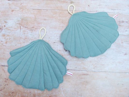 Ein Set aus zwei handgenähten Topflappen in der eleganten Form einer Muschel, gefertigt aus reinem, helltürkisem Leinen. Die Topflappen liegen flach nebeneinander auf einer hellen Küchenarbeitsplatte, sodass die geschwungene Muschelsilhouette gut sichtbar ist.