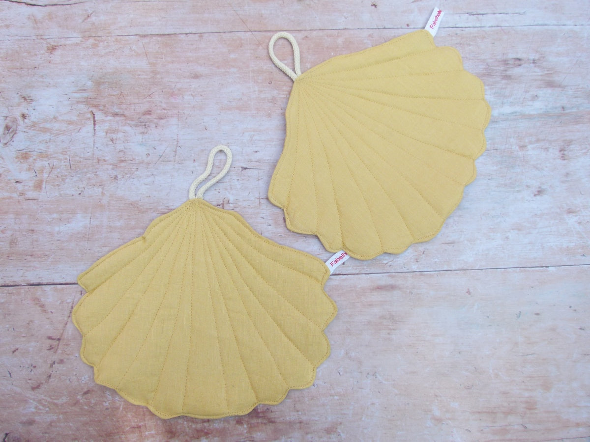 Ein Set aus zwei handgenähten Topflappen in der eleganten Form einer Muschel, gefertigt aus reinem, gelbem Leinen. Die Topflappen liegen flach nebeneinander auf einer hellen Küchenarbeitsplatte, sodass die geschwungene Muschelsilhouette gut sichtbar ist.