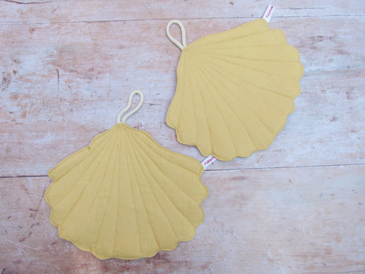 Ein Set aus zwei handgenähten Topflappen in der eleganten Form einer Muschel, gefertigt aus reinem, gelbem Leinen. Die Topflappen liegen flach nebeneinander auf einer hellen Küchenarbeitsplatte, sodass die geschwungene Muschelsilhouette gut sichtbar ist.