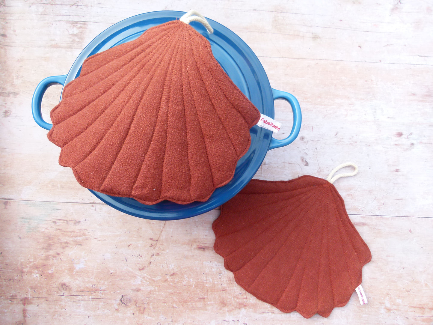 Präsentation des maritimen Topflappen-Sets an einem Kochtopf. Ein muschelförmiger Topflappen aus braunem Leinen liegt flach auf dem Topfdeckel, während der zweite Topflappen dekorativ daneben auf der Küchenzeile platziert ist.
