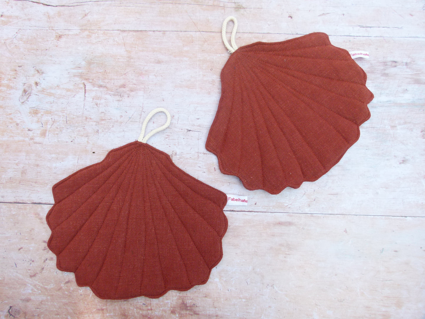 Ein Set aus zwei handgenähten Topflappen in der eleganten Form einer Muschel, gefertigt aus reinem, braunem Leinen. Die Topflappen liegen flach nebeneinander auf einer hellen Küchenarbeitsplatte, sodass die geschwungene Muschelsilhouette gut sichtbar ist.