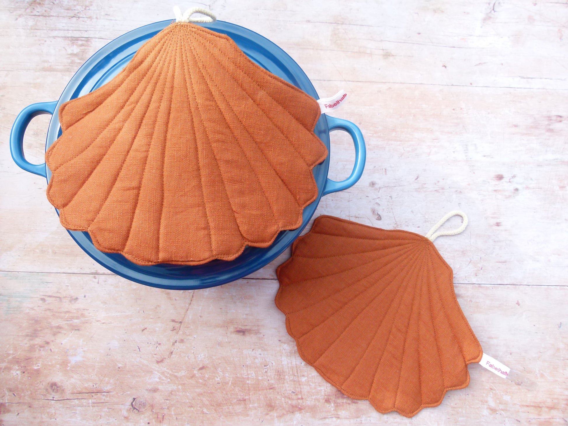 Präsentation des maritimen Topflappen-Sets an einem Kochtopf. Ein muschelförmiger Topflappen aus rostbraunem Leinen liegt flach auf dem Topfdeckel, während der zweite Topflappen dekorativ daneben auf der Küchenzeile platziert ist.