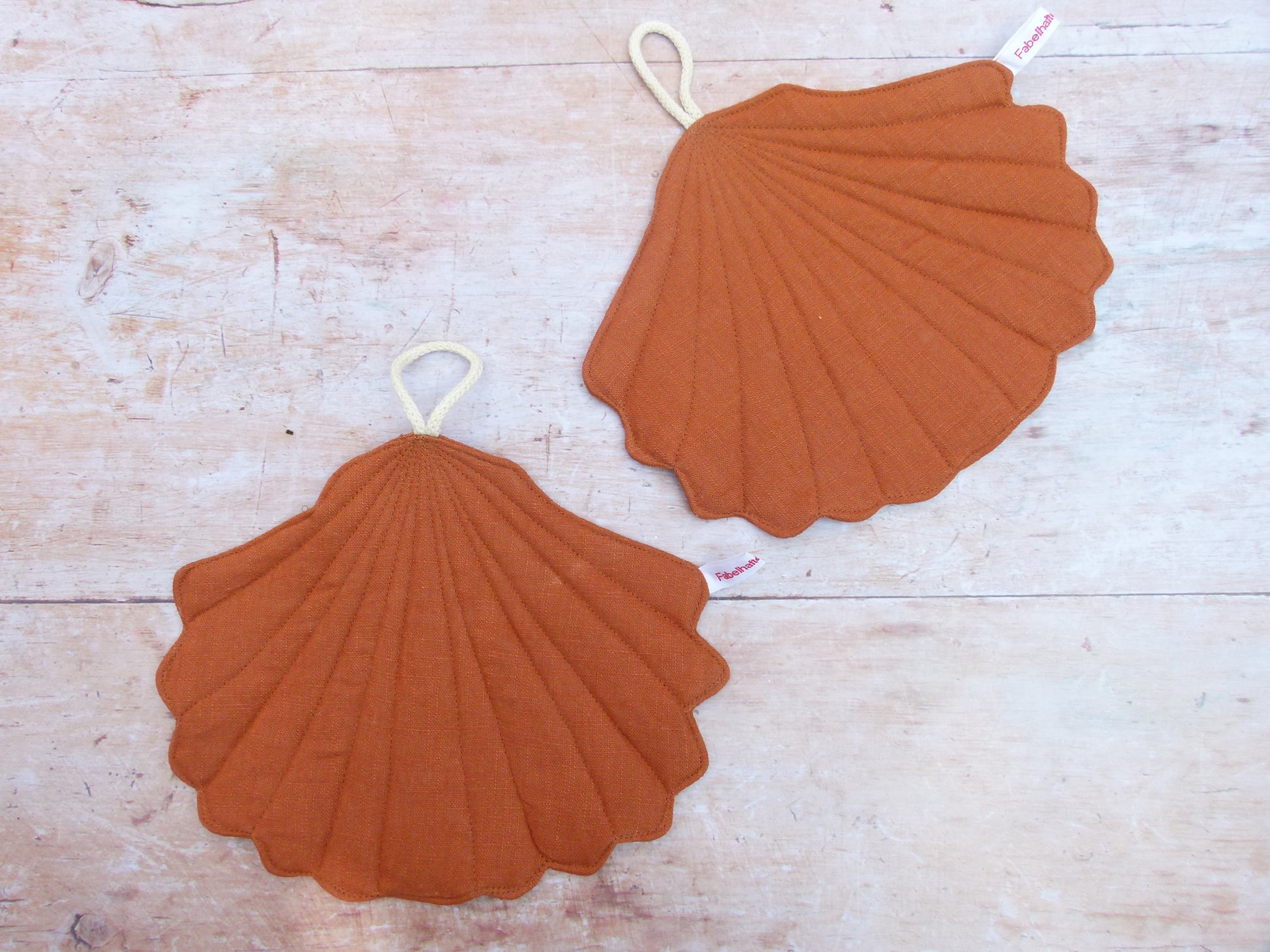 Ein Set aus zwei handgenähten Topflappen in der eleganten Form einer Muschel, gefertigt aus reinem, rostbraunem Leinen. Die Topflappen liegen flach nebeneinander auf einer hellen Küchenarbeitsplatte, sodass die geschwungene Muschelsilhouette gut sichtbar ist.