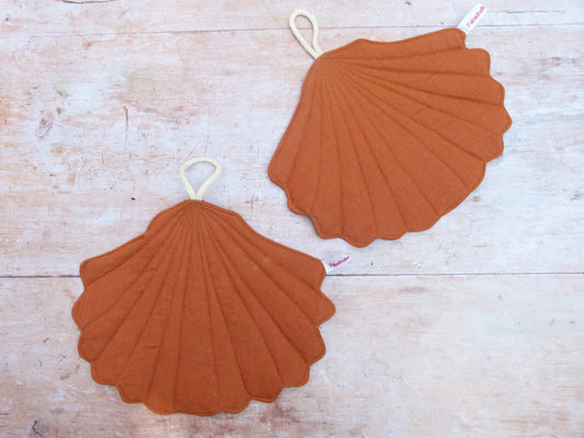 Ein Set aus zwei handgenähten Topflappen in der eleganten Form einer Muschel, gefertigt aus reinem, rostbraunem Leinen. Die Topflappen liegen flach nebeneinander auf einer hellen Küchenarbeitsplatte, sodass die geschwungene Muschelsilhouette gut sichtbar ist.
